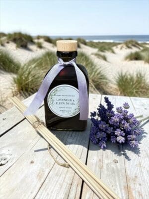 A bottle labeled “Lavender & Fleur de Sel Reed Diffuser” tied with a ribbon sits on a wooden table outdoors, accompanied by lavender flowers and diffuser sticks. Exuding serene sophistication, this luxury reed diffuser is set against sandy dunes and the ocean beneath a blue sky.