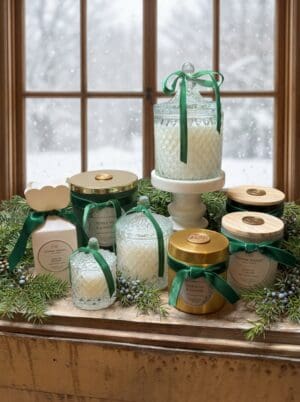 Assorted candles in decorative glass and metal jars with green ribbons are arranged on a wooden table accented with evergreen sprigs and complemented by the luxurious Noble Fir & Juniper Reed Diffuser, set in front of a snowy window.