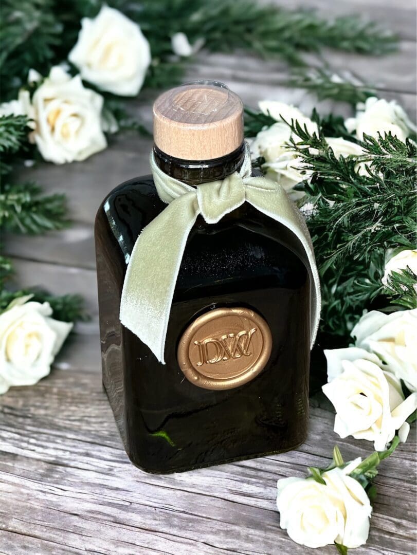 Donna Wright Designs French Rose & Evergreen Diffuser - 6 Oz: A dark glass bottle with a cork top, adorned with a green ribbon and a gold seal, is surrounded by white French roses and green foliage on a wooden surface.