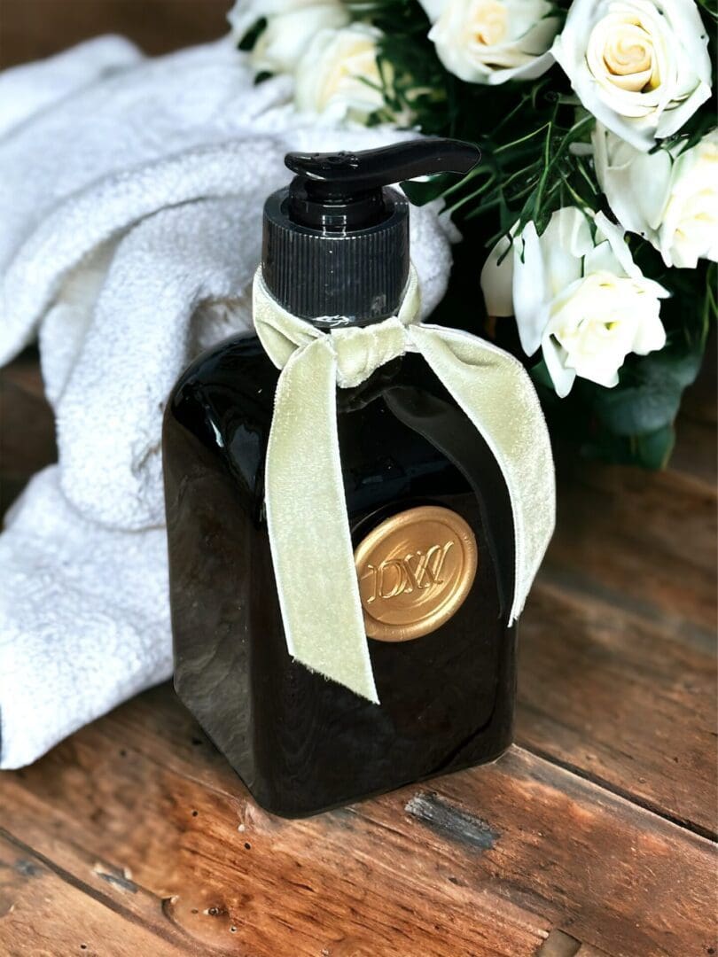 Donna Wright Designs A French Rose & Evergreen Hand Soap -8 Oz adorned with a gold label and a light beige ribbon, resting on a wooden surface next to white roses and a white towel.