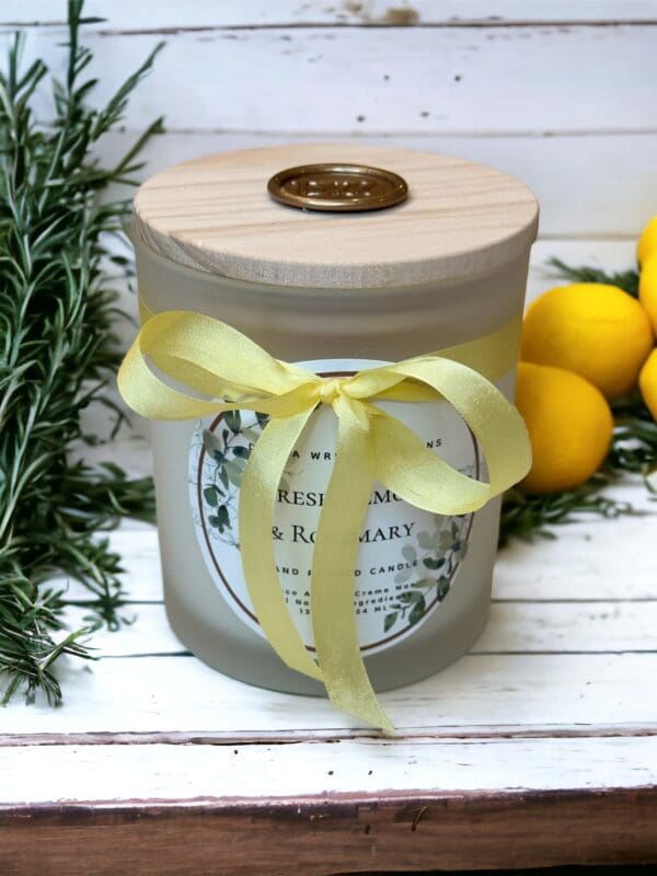 Fresh Lemon & Rosemary candle with wooden wicks 12 oz by Donna Wright Designs made of coco apricot creme wax and all natural ingredients & tied with a yellow ribbon. It's surrounded by fresh lemons and rosemary sprigs on a wooden table.