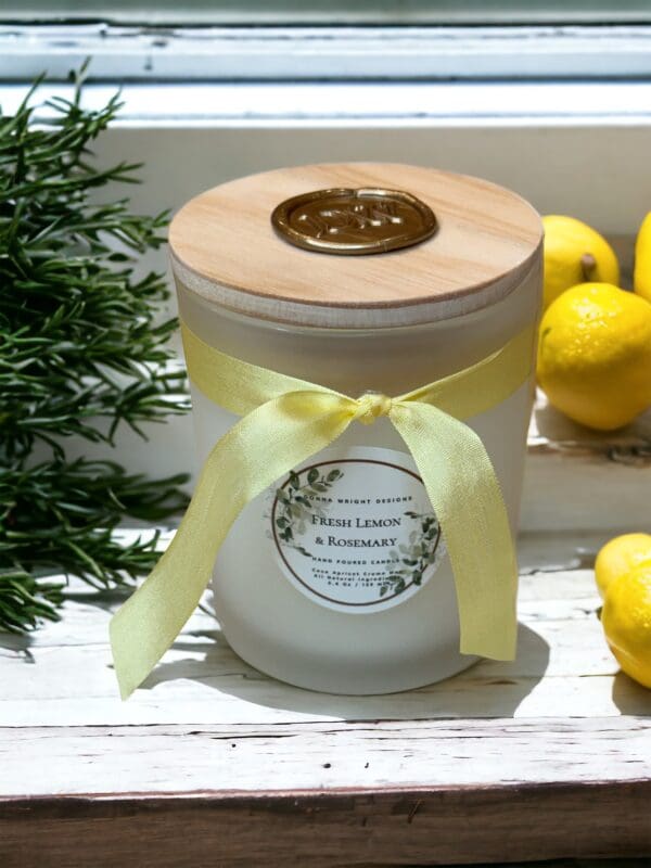 Fresh Lemon & Rosemary Candle 5.4 oz by Donna Wright Designs made with coco apricot creme wax & all natural ingredients, tied with a yellow ribbon, rests on a windowsill beside sprigs of rosemary and fresh lemons, illuminated by sunlight.