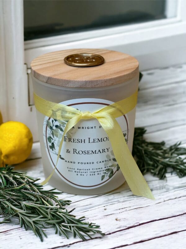 Fresh Lemon & Rosemary candle 8 oz by Donna Wright Designs, candle with wooden wick with a light wooden lid and yellow ribbon, made of coco apricot creme wax & all natural ingredients, set against a white wooden surface with fresh lemons and rosemary.