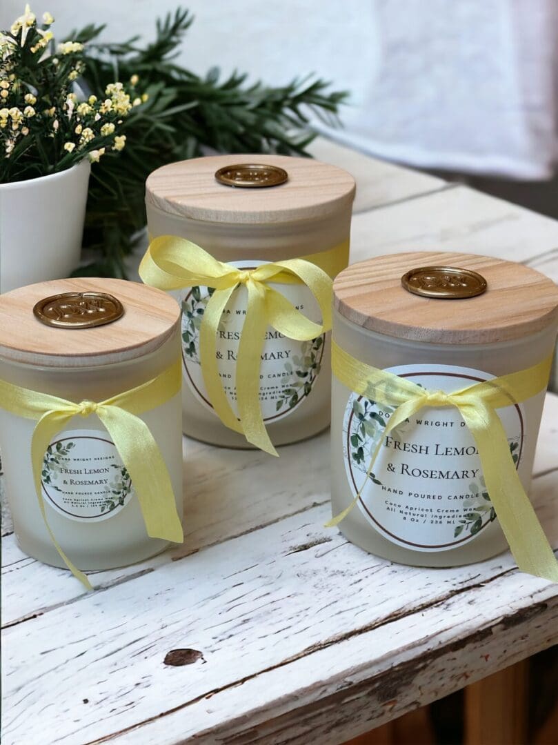 Three Fresh Lemon & Rosemary candles with wooden wicks by Donna Wright Designs in frosted glass vessels with yellow silk ribbon and wood lids sitting on a white wood table with yellow flowers & evergreen in the background.