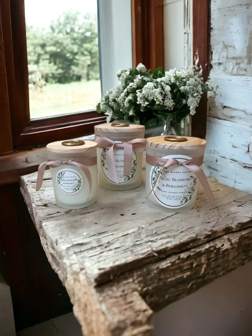 Donna Wright Designs Three lilac blossoms & bergamot candles on a rustic wooden table by a window overlooking a garden, with lilac blossoms in the background.