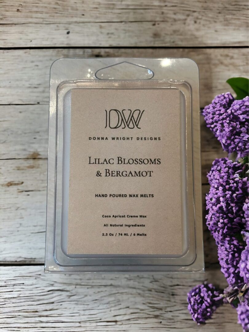 Donna Wright Designs Packaging of Lilac Blossoms & Bergamot Wax Melts with all-natural ingredients, displayed on a wooden surface next to purple lilac blossoms.