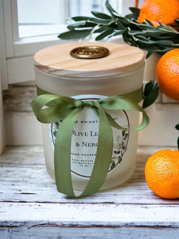 Olive Leaves & Neroli candle with wooden wick 12 oz by Donna Wright Designs in a frosted glass container with olive-colored silk ribbon and a wood lid on a white wood table with oranges & fresh olive tree branches