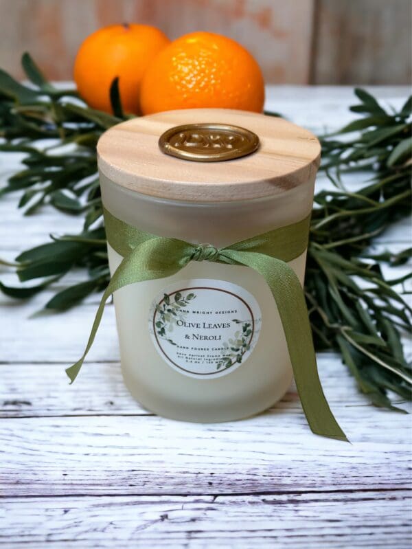 Olive Leaves & Neroli candle with wooden wick 5.4 oz by Donna Wright Designs in a frosted glass container with olive-colored silk ribbon and a wood lid on a white wood table with oranges & fresh olive tree branches