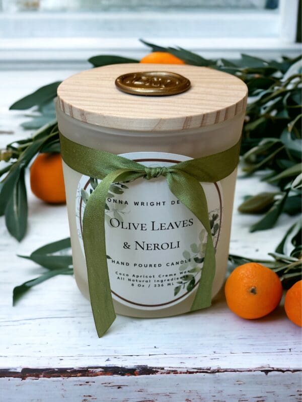 Olive Leaves & Neroli candle with wooden wick 8 oz by Donna Wright Designs in a frosted glass container with olive-colored silk ribbon and a wood lid on a white wood table with oranges & fresh olive tree branches