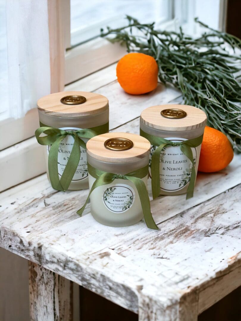 Donna Wright Designs Three jars of Olive Leaves & Neroli Candles with wooden lids, tied with green ribbons, situated on a distressed white wooden table alongside fresh oranges and olive leaves.