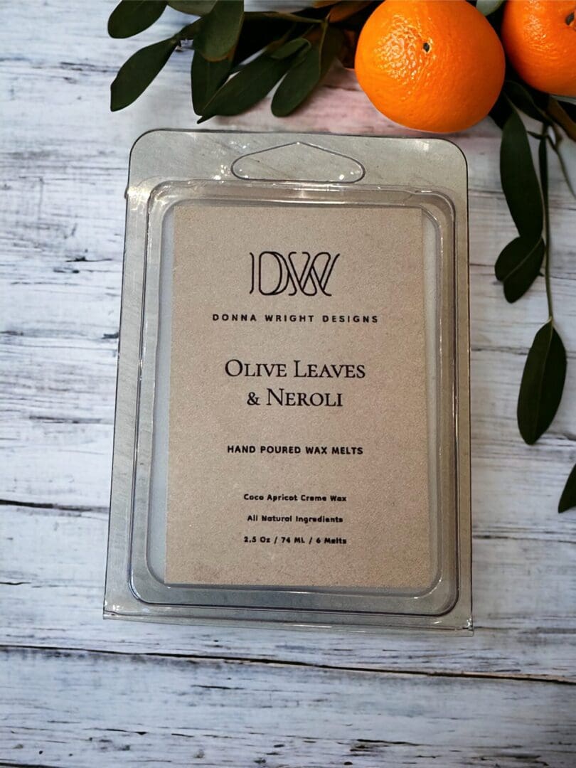 Olive Leaves & Neroli wax melts 2.5 oz from Donna Wright Designs with perfume grade fragrance & essential oils in a clear plastic package with 6 melts on a white wood table with oranges & olives leaves in the background.