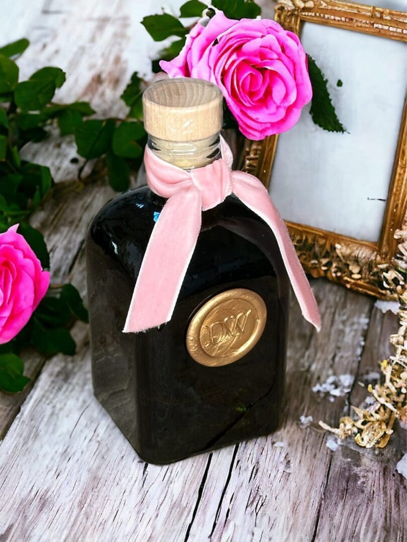 Donna Wright Designs Spring Bouquet Diffuser - 6 Oz with a pink ribbon tied around its neck, bearing a gold seal, sits on a wooden surface next to French Rose roses and a vintage gold frame.