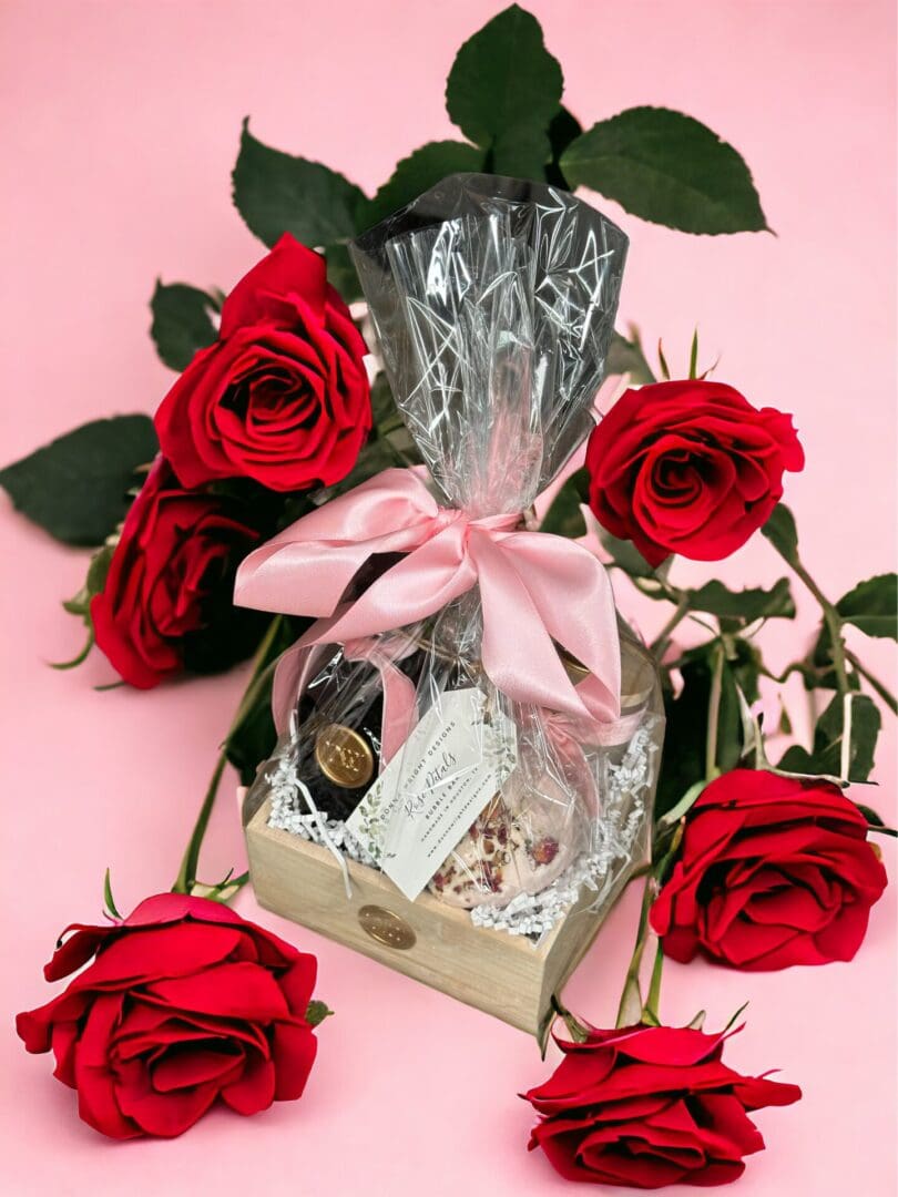 Donna Wright Designs A Spring Bouquet - Medium Gift Basket wrapped in clear cellophane with a pink ribbon, surrounded by vibrant red roses on a soft pink background. The basket contains items, partly visible, with a classy, festive.