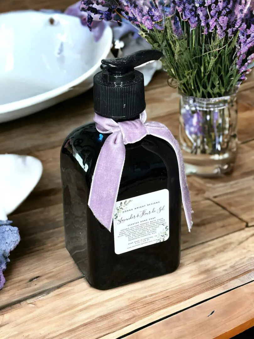 Lavender scented hand soap by Donna Wright Designs in Lavender & Fleur de Sel 8 oz olive-colored bottle with purple ribbon on a wood desk with fresh lavender in the background.