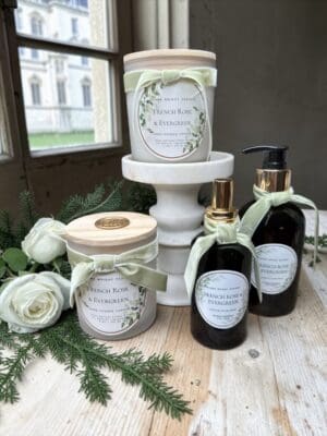 Two pump bottles of French Rose & Evergreen Hand Soap and two scented candles in glass jars, all labeled “French Rose & Evergreen,” are arranged on a wooden table with greenery and white roses near a window.