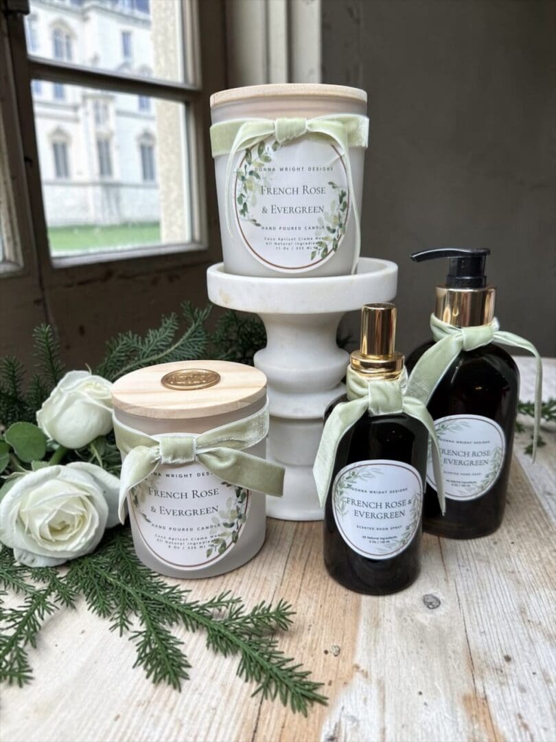 Two pump bottles of French Rose & Evergreen Hand Soap and two scented candles in glass jars, all labeled “French Rose & Evergreen,” are arranged on a wooden table with greenery and white roses near a window.