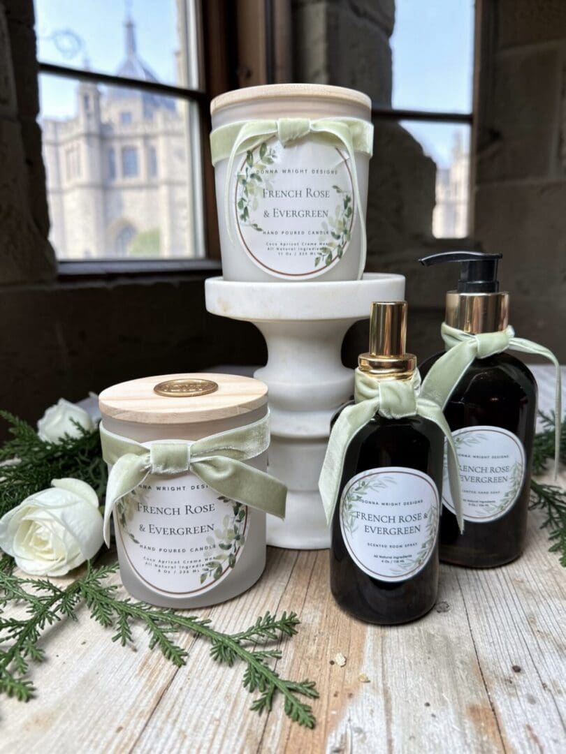 French Rose & Evergreen Hand Soap on a white stand, decorated with green bows, set on a rustic table with evergreens; stone building and window in the background.