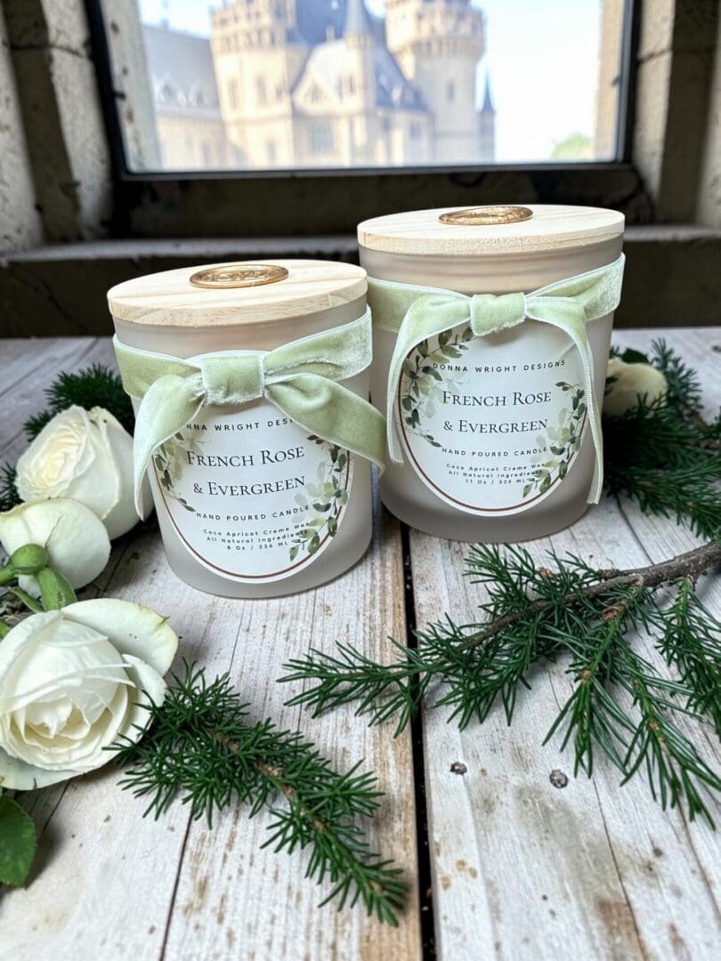 Two white candles labeled “French Rose & Evergreen” rest on a stone ledge alongside pine sprigs and white roses, capturing the refined essence of the French Rose & Evergreen Hand Soap. Light green ribbons and wooden lids accent each candle for an elegant touch, while a blurred castle appears outside the window in the background.