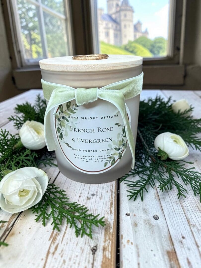 A “French Rose & Evergreen” candle tied with a green ribbon rests on a rustic white table, surrounded by evergreen branches and white flowers. Next to it is a bottle of French Rose & Evergreen Hand Soap, all set in front of a window overlooking a castle.