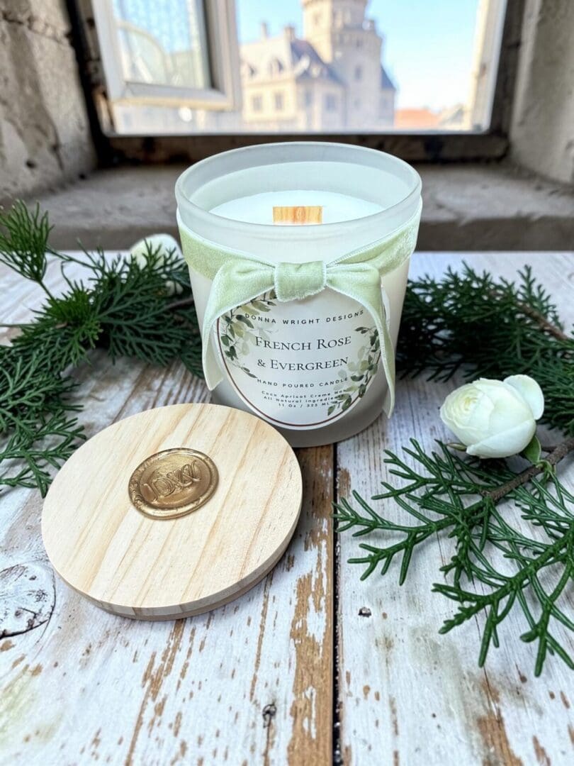 A bottle of French Rose & Evergreen Hand Soap sits on a wooden surface by a window, surrounded by lush greenery, a white flower, and its elegant wooden lid with a gold seal. Reminiscent of refined French luxury, this scene imbues your space with an air of sophistication as a building stands visible through the window.