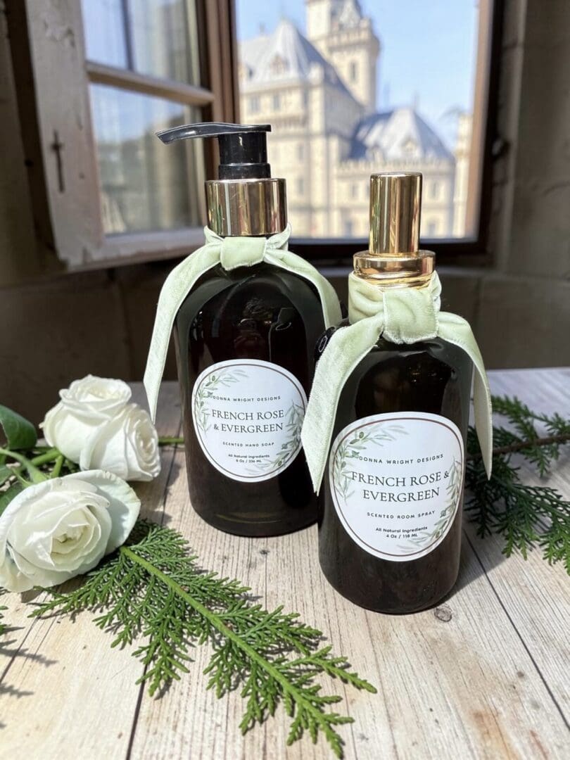 Two dark pump bottles labeled “French Rose & Evergreen Hand Soap,” each adorned with green ribbon bows, sit on a table surrounded by white roses and evergreen branches. Sunlight streams through a window, illuminating the elegant display against the backdrop of a historic building.