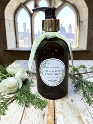 A bottle of French Rose & Evergreen Hand Soap, adorned with a light green bow, rests on a wooden surface surrounded by lush greenery inside a building with arched windows that offer views of the city skyline.