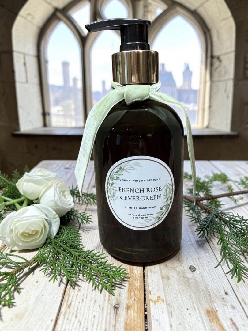 A bottle of French Rose & Evergreen Hand Soap, adorned with a light green bow, rests on a wooden surface surrounded by lush greenery inside a building with arched windows that offer views of the city skyline.
