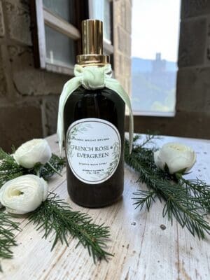 A bottle of French Rose & Evergreen room spray with a bow sits on a rustic table, surrounded by white flowers and evergreen sprigs. Nearby, the elegant packaging of the French Rose & Evergreen Hand Soap adds to the sophisticated ambiance, with a window and blurred outdoor scenery in the background.