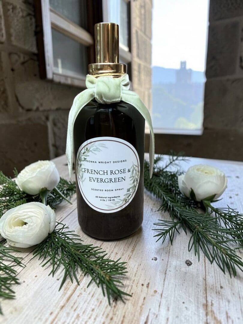 A bottle of French Rose & Evergreen room spray with a bow sits on a rustic table, surrounded by white flowers and evergreen sprigs. Nearby, the elegant packaging of the French Rose & Evergreen Hand Soap adds to the sophisticated ambiance, with a window and blurred outdoor scenery in the background.