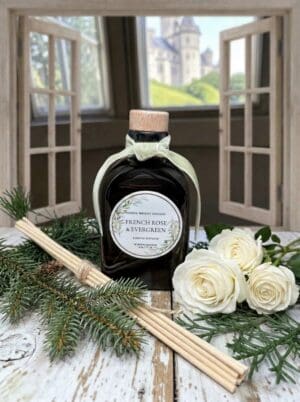 The French Rose & Evergreen Reed Diffuser, featuring elegant reed sticks, white roses, and evergreen sprigs, rests on a rustic table by an open window with a distant castle in view.