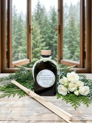 The French Rose & Evergreen Reed Diffuser is displayed on a wooden table alongside a bouquet of white roses and its accompanying reed sticks. Evergreen branches surround the diffuser, and through a window in the background, green trees can be seen outside.