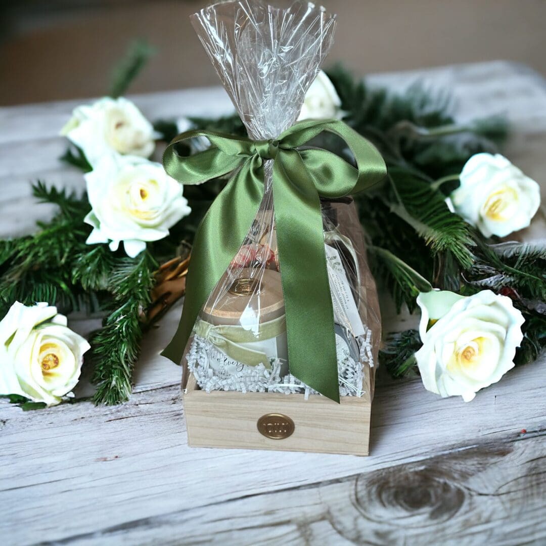 Donna Wright Designs A French Rose & Evergreen - Medium Candle Gift Basket featuring scented candles in a transparent cone wrapped in cellophane and tied with a green satin ribbon, placed in a wooden box surrounded by white roses and green fern leaves.