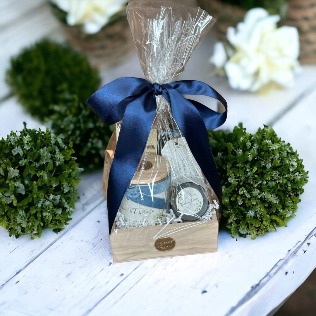 Donna Wright Designs A gift-wrapped clear bag containing a Gardenia & Tuberose - Medium Gift Basket and a message scroll, tied with a dark blue ribbon, presented in a wooden box surrounded by gardenia bushes on a white wooden surface.