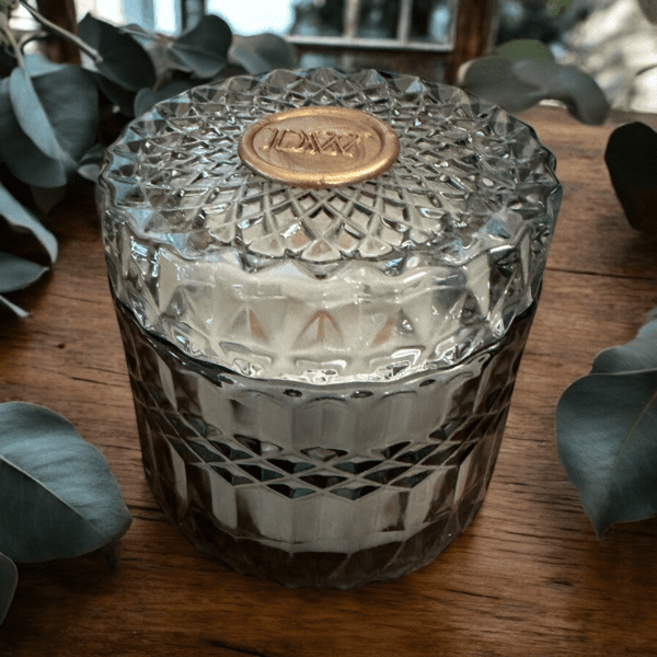 A Eucalyptus & Garden Mint candle in a blue glass container by Donna Wright Designs sitting on a wood table with eucalyptus behind it