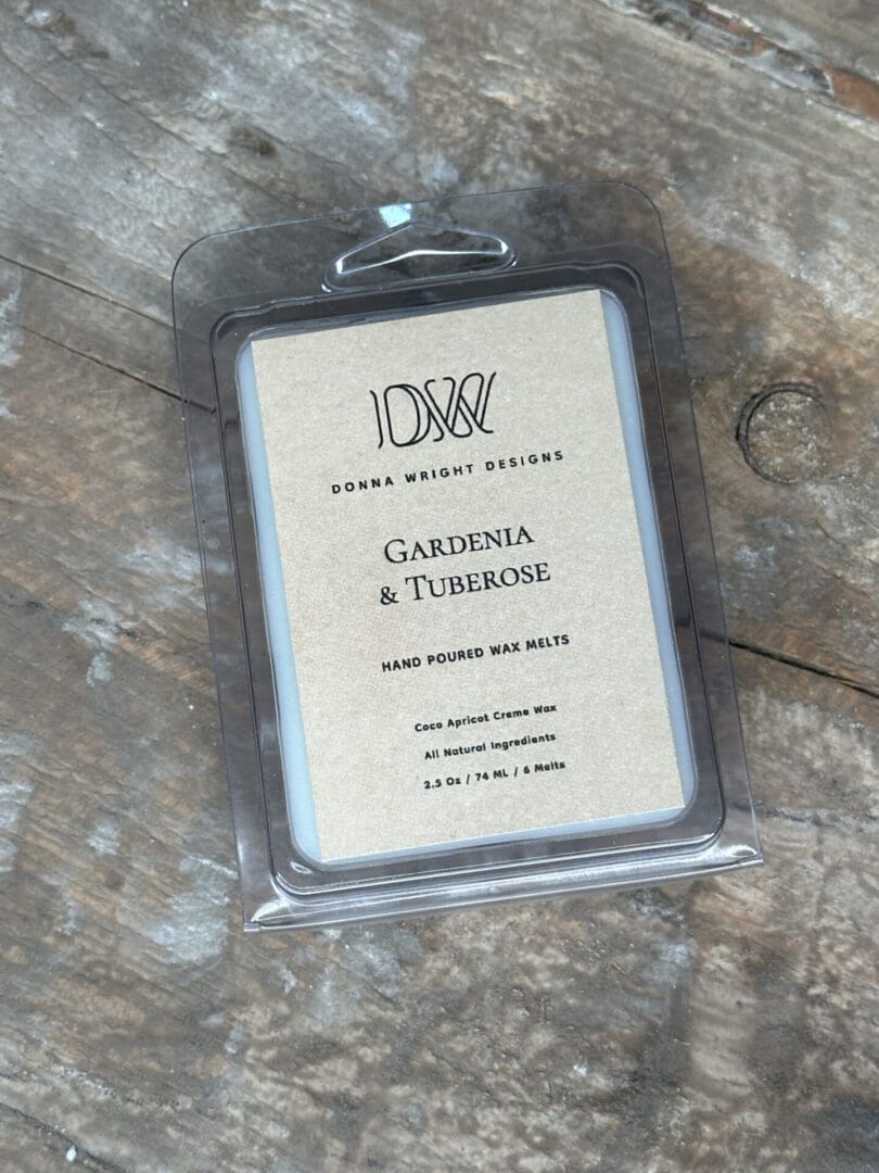 A Donna Wright Designs Gardenia & Tuberose wax melts package in transparent plastic with a brown label listing the product details, and the wax is visible. It's placed on a rustic wooden surface.