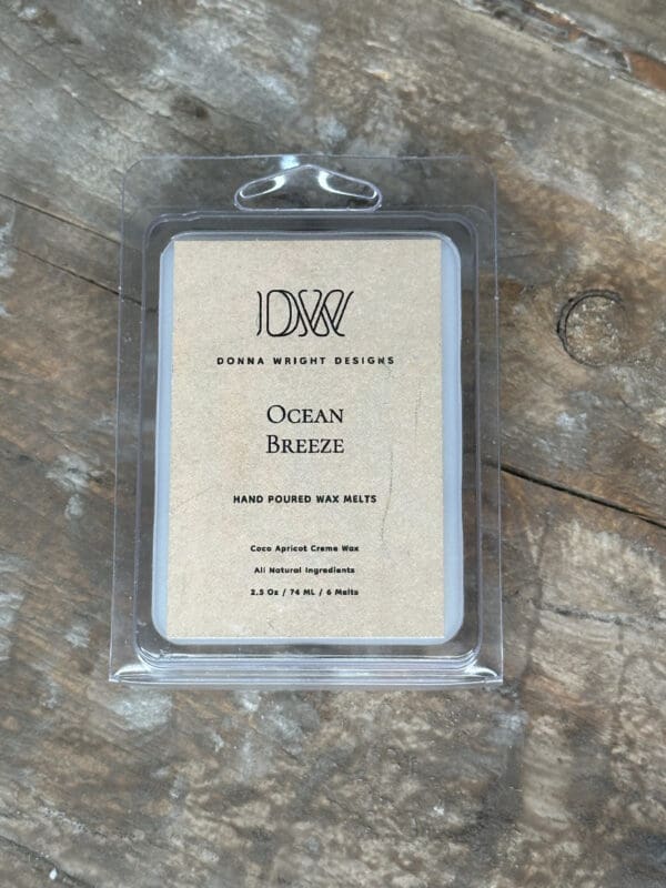 Donna Wright Designs A packaged set of "Ocean Breeze" hand-poured wax melts from Donna Wright Designs is displayed on a wooden surface. The label indicates the product is made of coconut apricot cream wax, weighs 2.5 oz, and contains 6 melts with all-natural ingredients.