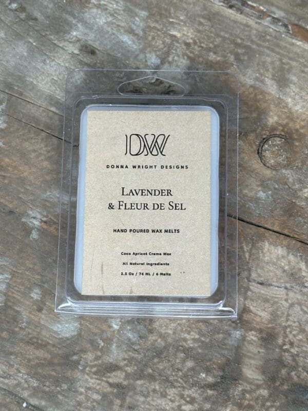 A package of lavender and fleur de sel scented wax melts from Donna Wright Designs is displayed. The package is made of clear plastic, showing six individual wax melts inside. The label indicates it's made with 100% all-natural coco apricot creme wax. The background is a rustic wooden surface.