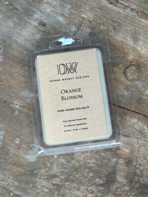 Donna Wright Designs A clamshell package of wax melts labeled "Donna Wright Designs - Orange Blossom" is placed on a rustic wooden surface. The label notes that the product is hand-poured, made with natural ingredients, and cocoa-apricot crème wax. The package contains six wax melts.