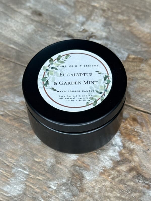 A small black tin candle container with a white label on the lid that reads, "Eucalyptus & Garden Mint" and "Hand Poured Candle." The label also features a decorative wreath of green leaves. The container is placed on a rustic wooden surface.