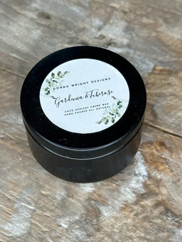 Donna Wright Designs A black jar sits on a wooden surface. The jar's lid features a white label with green foliage detailing. The label reads: "Donna Wright Designs, Gerlavia & Therese, Coco Apricot Cream Wax, Hand Poured All Natural.