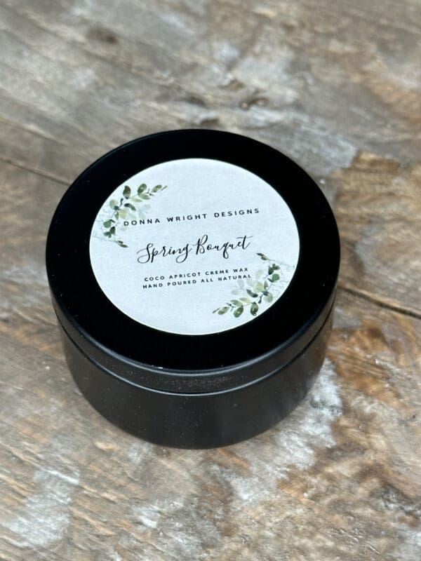 Donna Wright Designs A black metal tin with a lid labeled "Donna Wright Designs" and "Spring Bouquet" on a wooden surface. The label also notes that it contains a coco apricot crème wax and is hand-poured and all-natural. The design includes green botanical illustrations.