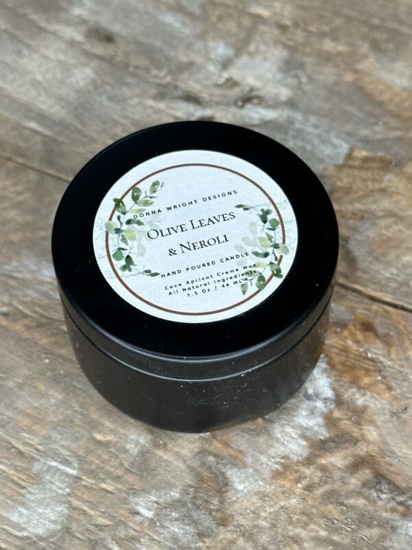 A round, black tin of "Olive Leaves & Neroli" hand-poured candle sits on a rustic wooden surface. The lid features a white label with decorative green leaves and text detailing the candle's fragrance and brand, "Donna Wright Designs.