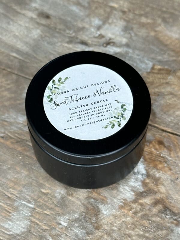 Donna Wright Designs A black round tin containing a scented candle with the lid on. The label on the lid reads "Bonnie Wright Designs, Sweet Tobacco & Vanilla, Scented Candle, Eco-friendly, hand-poured in Idaho" along with a website address and green leafy illustrations around the text.