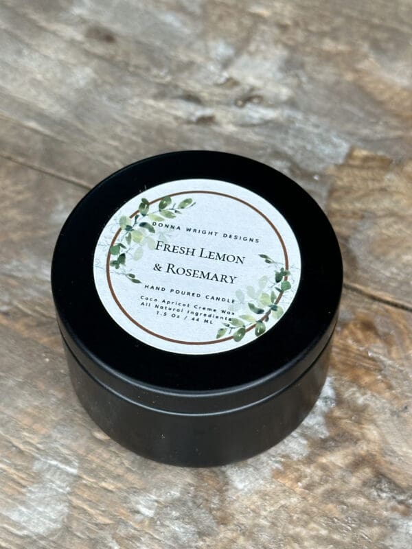 A small black tin container with a circular label on top placed on a rustic wooden surface. The label reads, "Fresh Lemon & Rosemary, Hand Poured Candle," and features a decorative green leaf design around the edges.