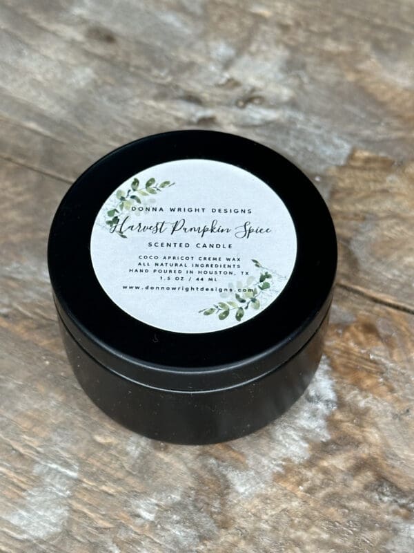 Donna Wright Designs A black tin container sits on a wooden surface. The lid features a white label with botanical accents and text: "Donna Wright Designs - Harvest Pumpkin Spice - Scented Candle - Coco Apricot Crème Wax - All Natural Ingredients - Hand Poured in Brooklyn, NY - 1.5 oz / 42 ml.