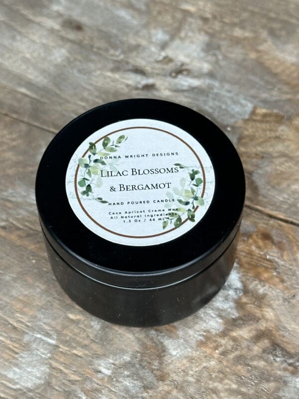 Donna Wright Designs A black cylindrical candle tin with a white label on top sits on a wooden surface. The label reads "Lilac Blossoms & Bergamot" and "Hand Poured Candle," adorned with an illustration of green leaves. The background is a rustic wooden table.