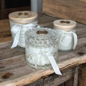 Three Woodland Lily & Vetiver Grass candles by Donna Wright Designs in decorative jars sit on a rustic wooden surface. The two in the back have frosted glass jars with wooden lids and white labels, tied with white ribbons. The one in the front has a clear, intricately-patterned glass jar with a white ribbon.