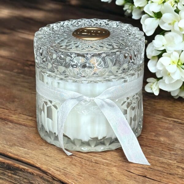 Donna Wright Designs A decorative, transparent glass jar with an intricate pattern holds a white candle inside. The jar is adorned with a pale ribbon tied in a bow around its middle and features a round, golden emblem on the lid. White flowers are blurred in the background on a wooden surface.