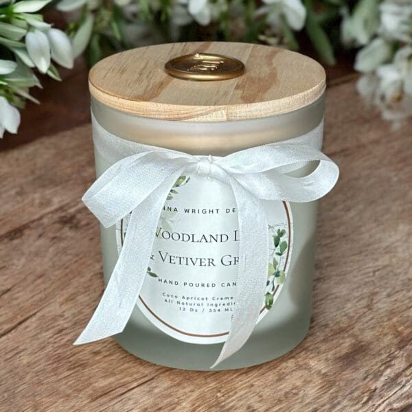 A glass candle jar with a light wooden lid, adorned with a white ribbon bow. The label reads "Woodland Lily & Vetiver Grass" and mentions it is a hand-poured candle. The scene includes a wooden surface and some blurred white flowers in the background.