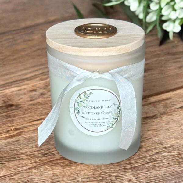 A frosted glass candle with a wooden lid is adorned with a white ribbon bow and a wax seal. The label reads “Woodland Lily & Vetiver Grass,” and floral illustrations decorate the sides. The candle is placed on a wooden surface with greenery in the background.